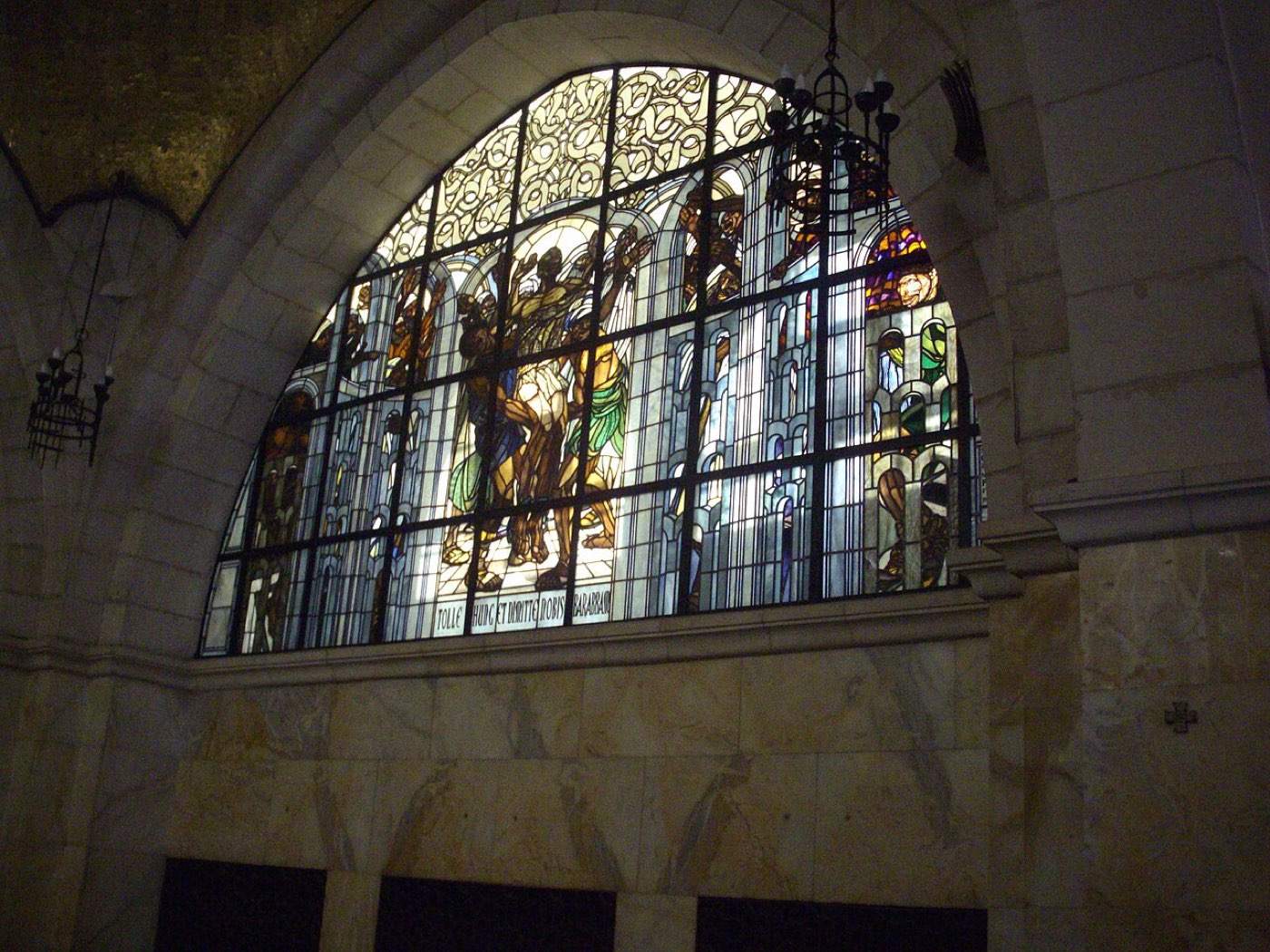A stained-glass window of the release of Barabbas: Pilate on his tribunal gestures between Jesus and the prisoner as the crowd chooses, with a Latin scroll reading Tolle hunc et dimitte nobis Barabbam.