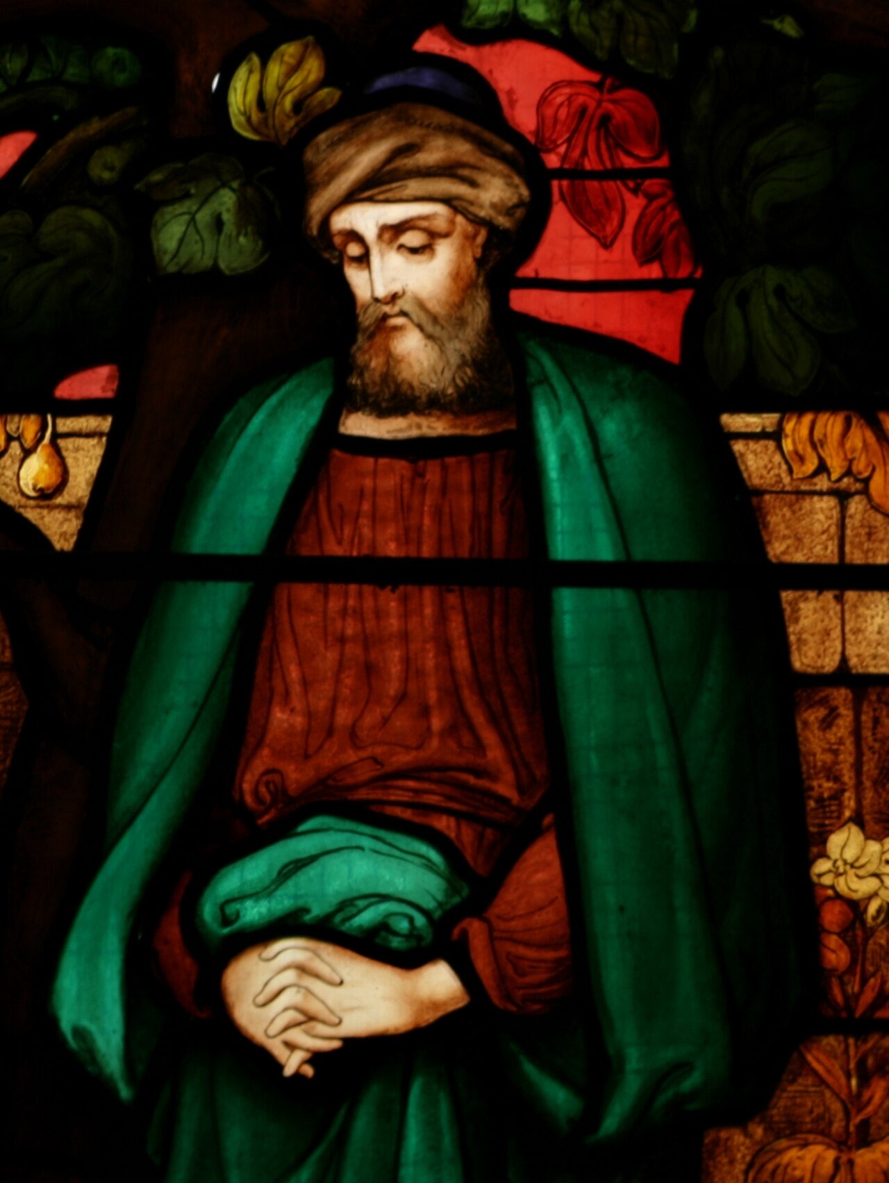 A stained-glass window of Nathanael: a bearded man in a red tunic and green cloak stands with hands folded in thought beneath the leaves of a fig tree.