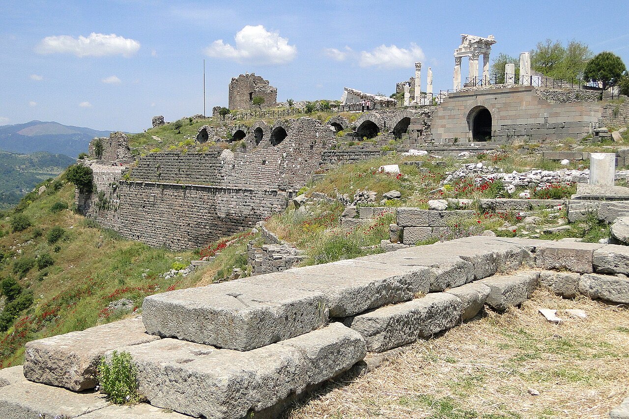 The hilltop acropolis of Pergamon: massive terrace walls, arched foundations, and the white columns of the Trajan temple against green mountains.