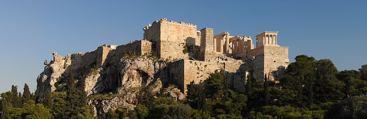 Panorama of the Acropolis of Athens from the Areopagus hill: fortress walls, the Propylaea gates, and temple columns in evening light above wooded slopes.