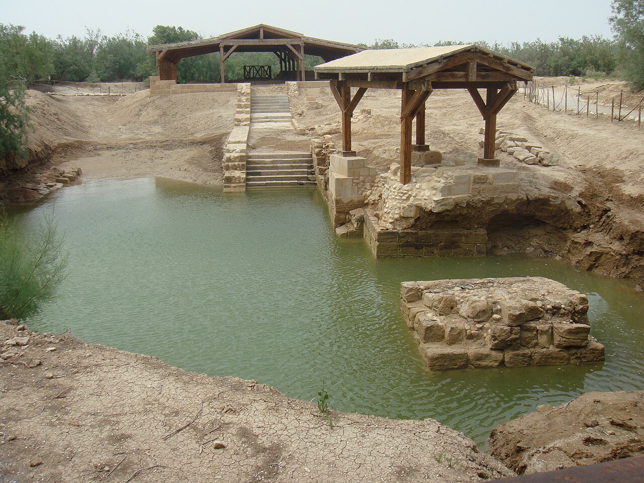 Excavated stone steps descending into a spring-fed green pool at the Al-Maghtas baptism site, with modern wooden shelters protecting the ruins.