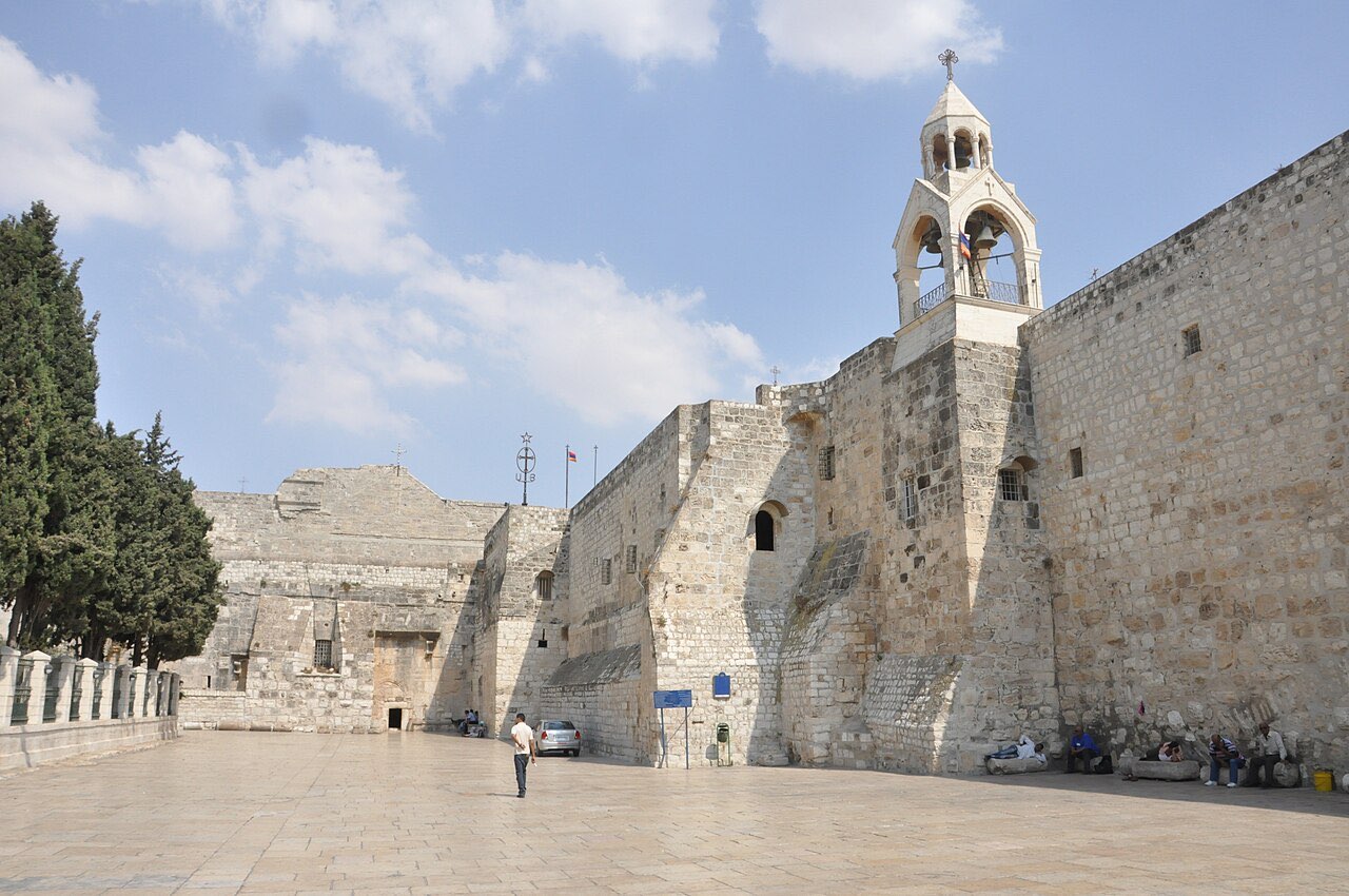The fortress-like stone facade of the Church of the Nativity across a wide paved square, a bell tower rising on the right and a lone person crossing the pavement.