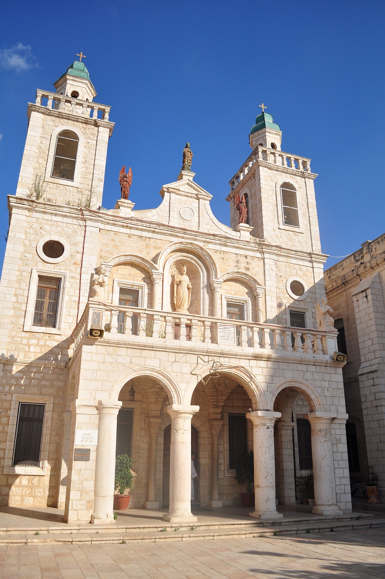 The Franciscan Wedding Church at Kafr Kanna: a pale stone facade with twin towers capped in green domes, statues above the balustrade, against a deep blue sky.