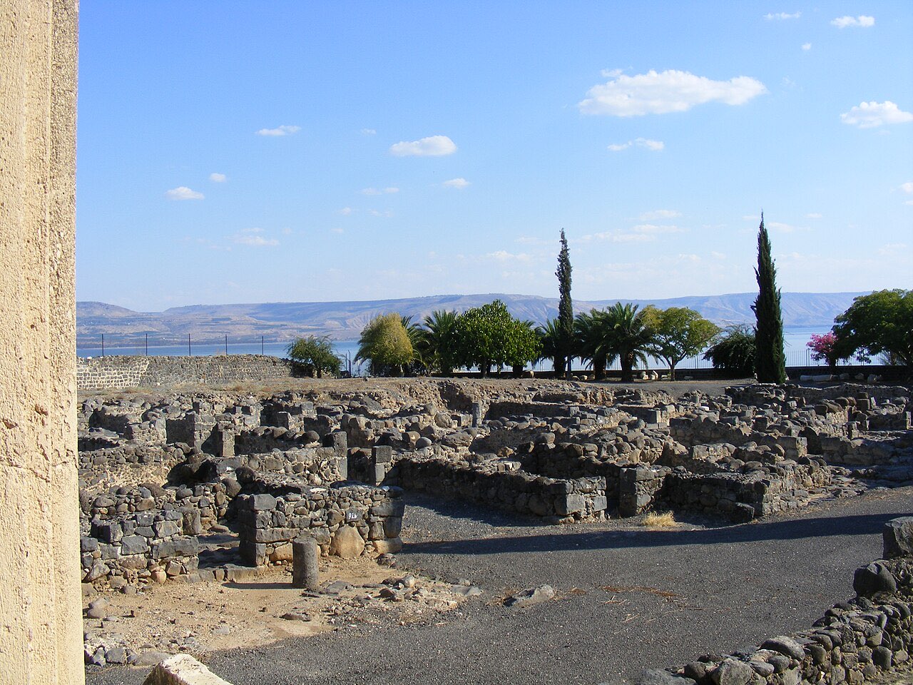 Excavated black basalt walls of ancient Capernaum's houses in the foreground, with palms and cypresses behind them and the Sea of Galilee and far hills in the distance.