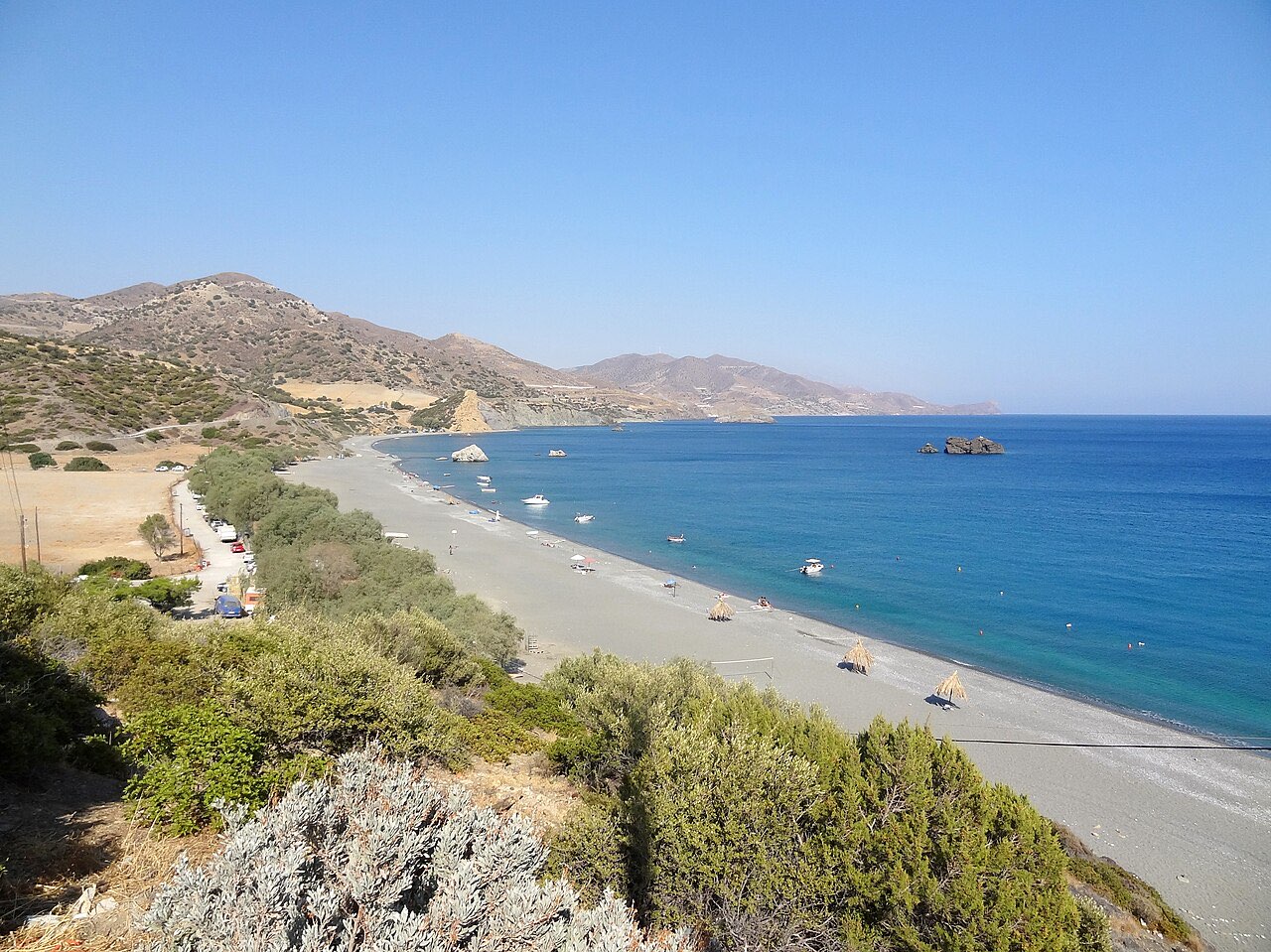 The curved bay of Kali Limenes on Crete's south coast: a gravel beach, low green scrub, brown hills, and small rocky islands in a deep blue sea.