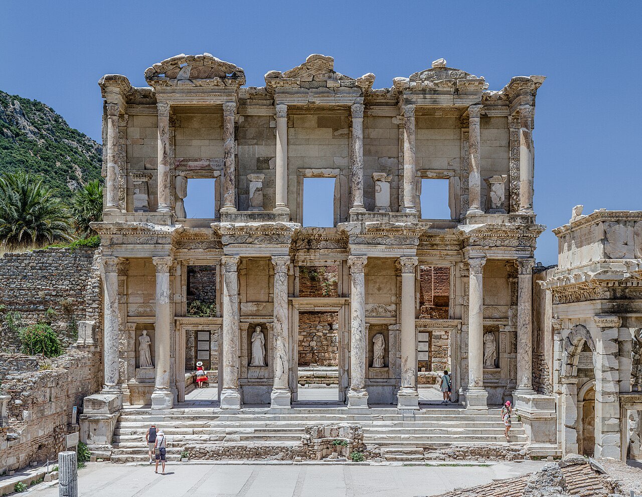 The two-story columned facade of the Library of Celsus at Ephesus, with statues in its niches and tourists small at its base.