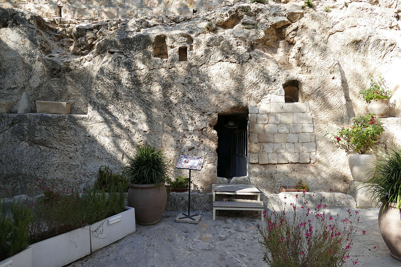 A rock face in a quiet garden with a rectangular tomb doorway cut into it and a channel for a rolling stone at its base.