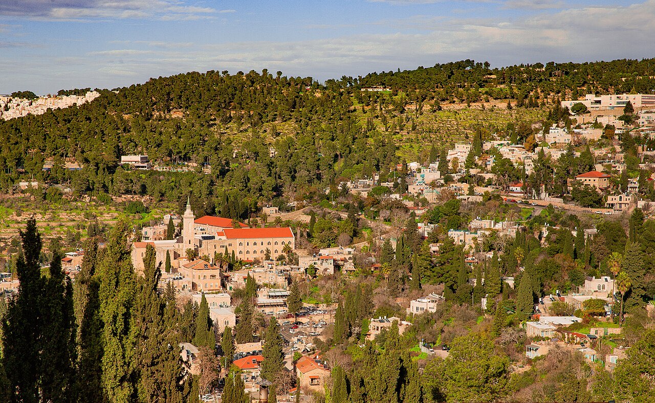 The village of Ein Karem on a terraced hillside: stone houses among cypress and pine trees, with a red-roofed church and its tower rising at the center.