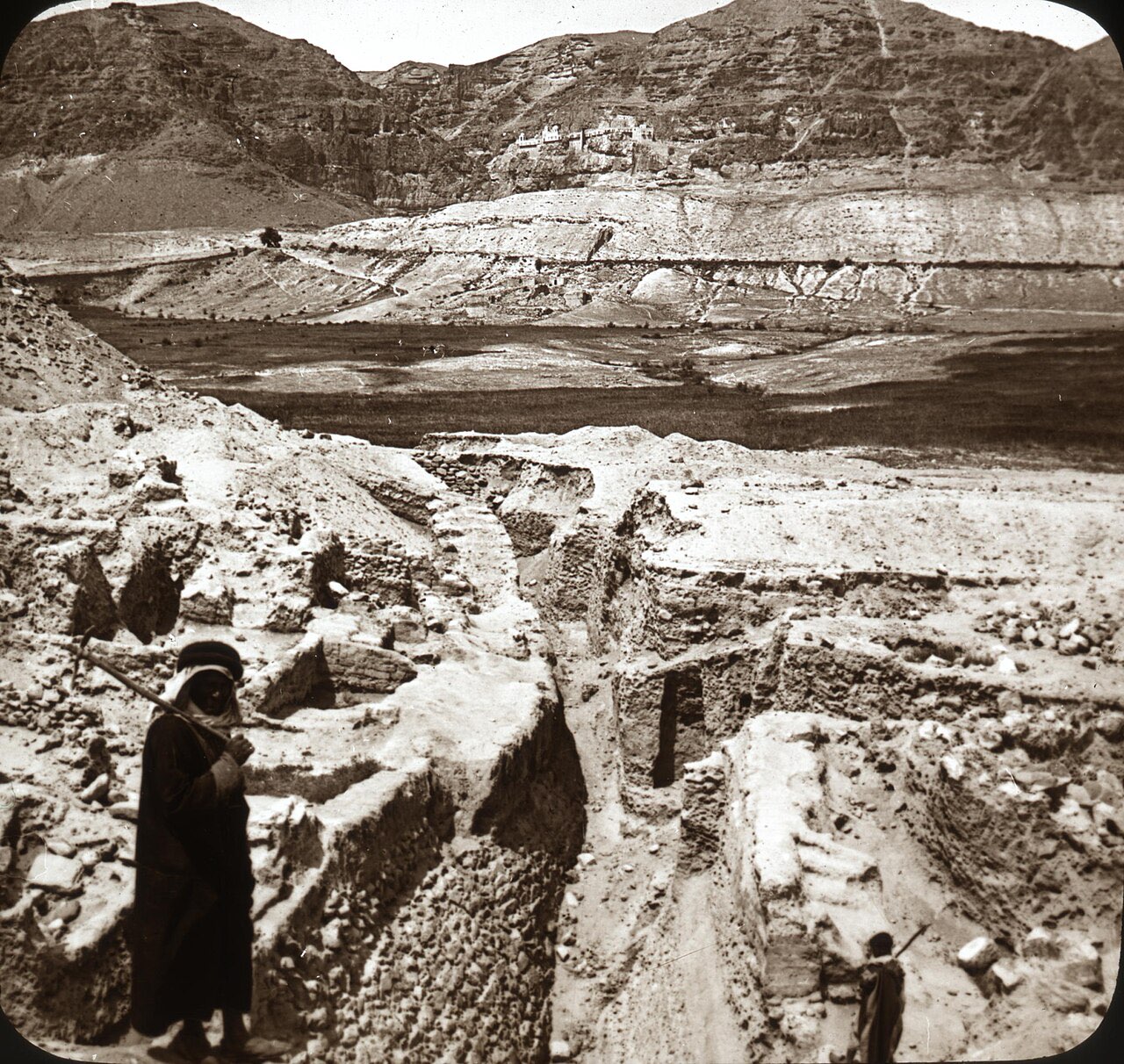 An early photograph of the Jericho excavations: a deep trench through the ancient mound, with the cliffs of the Mount of Temptation and its clinging monastery rising behind.