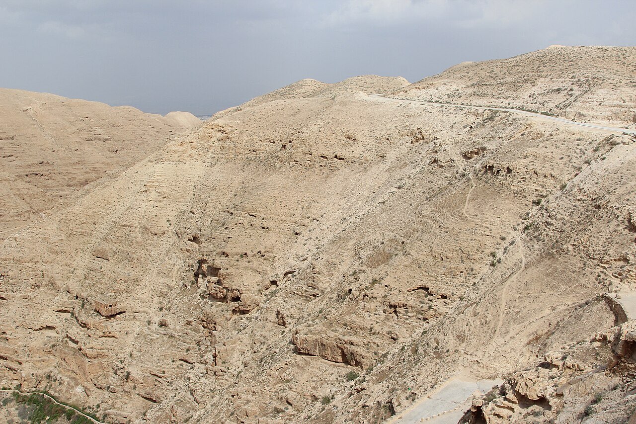 Bare, sun-bleached hills of the Judean wilderness cut by the deep ravine of Wadi Qelt, with a thin footpath tracing the slope and no building in sight.
