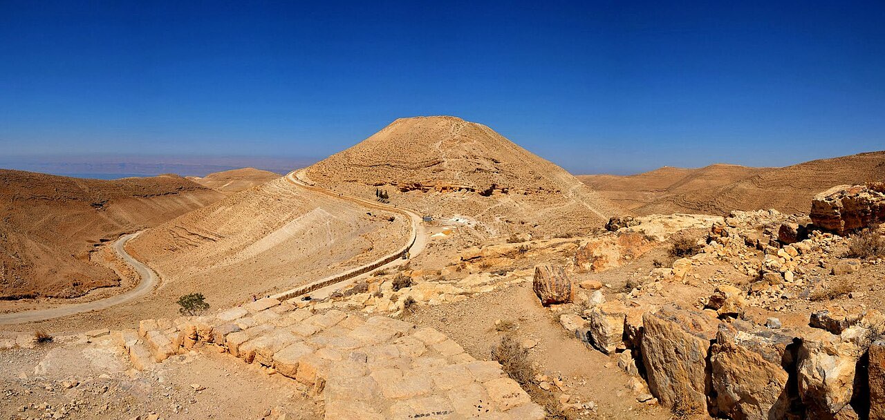 The conical hilltop of Machaerus crowned with fortress ruins, standing alone above deep desert ravines, with the Dead Sea hazy in the distance.