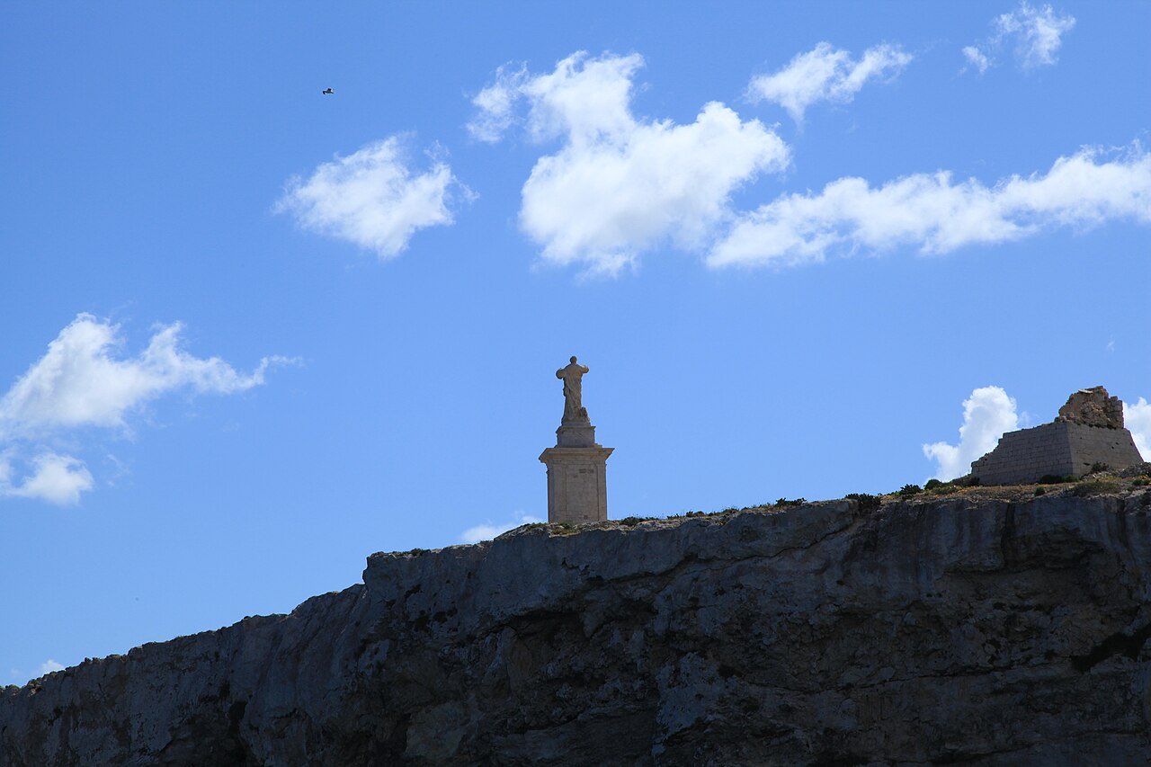 The white statue of St. Paul on its pedestal atop the rocky islet at St. Paul's Bay, Malta, against a blue sky.
