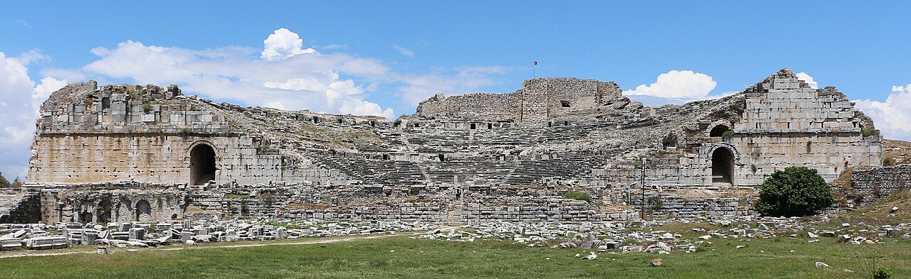 The wide stone sweep of the ancient theatre of Miletus, its tiers of seats rising to ruined upper galleries under a bright sky.