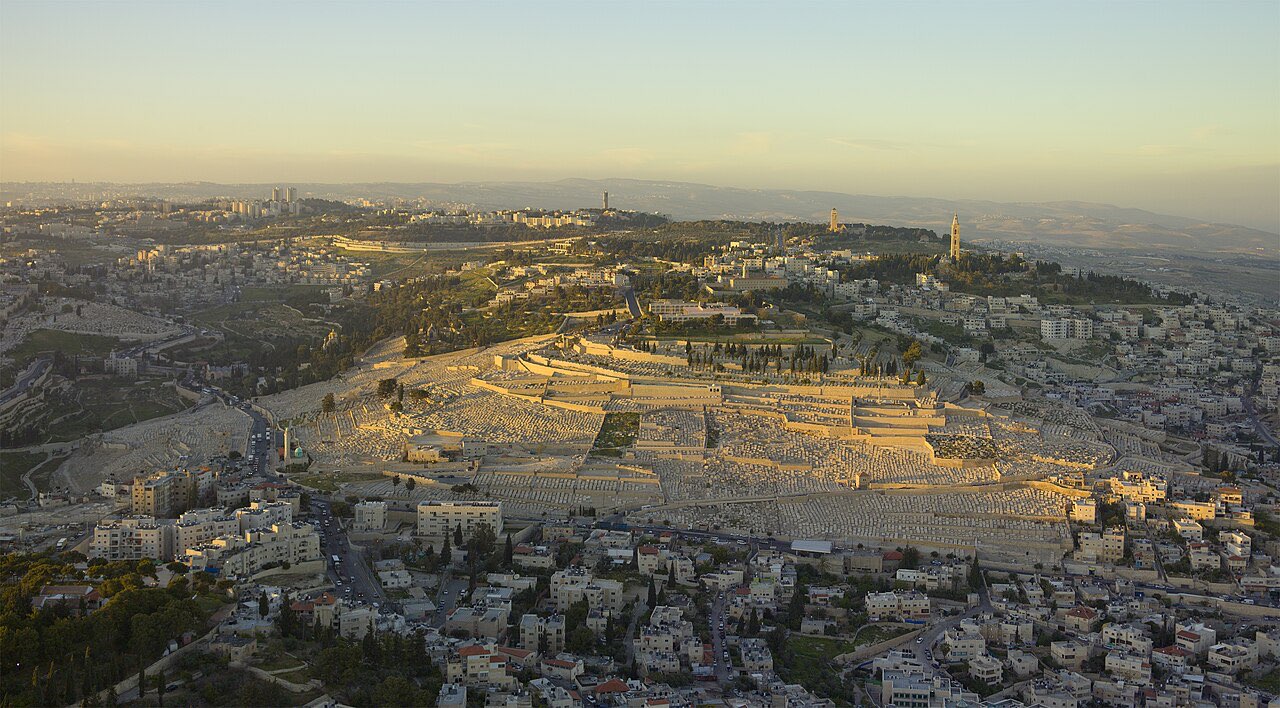 Aerial view of the Mount of Olives at golden hour: the vast terraced Jewish cemetery on the slope, church towers on the ridge, and the Kidron valley below.