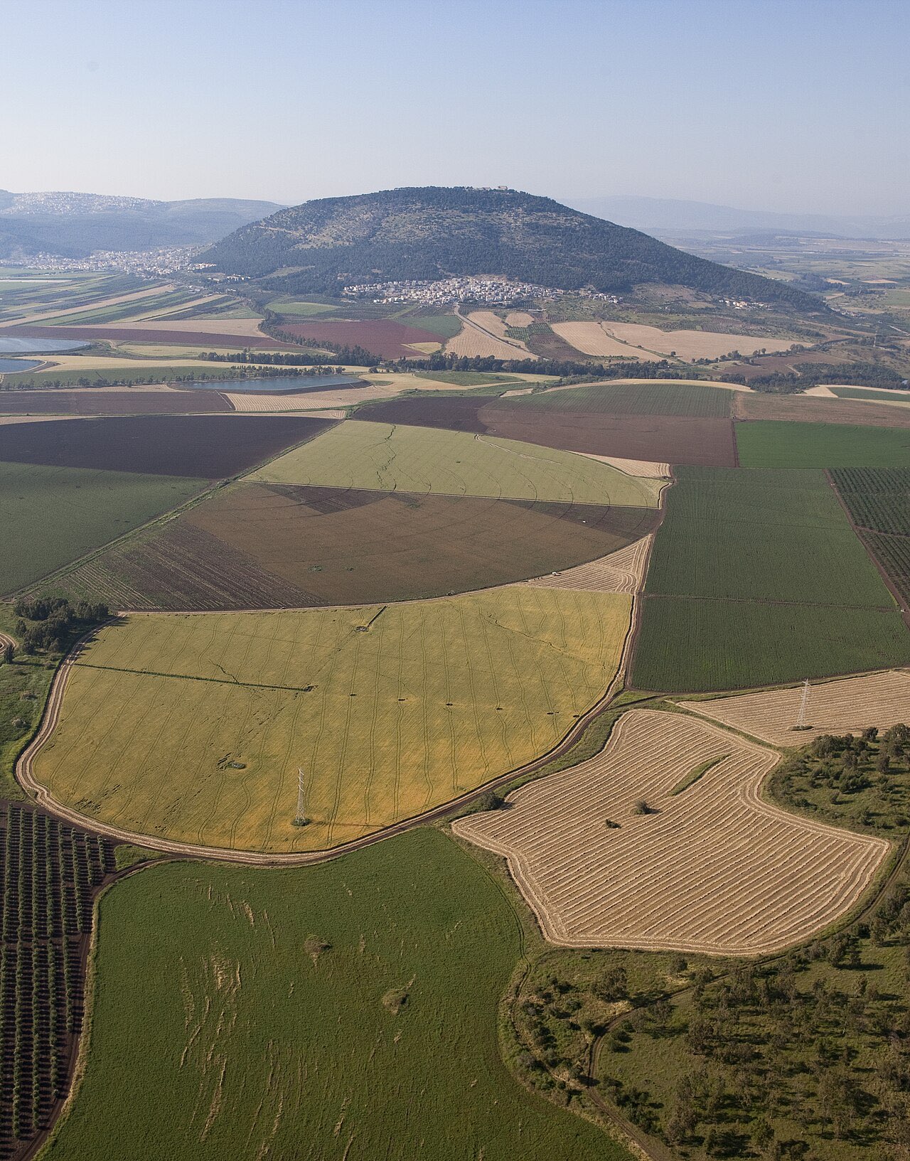 Aerial view of Mount Tabor: a solitary dome-shaped mountain rising from a patchwork of farmland, its summit ringed by trees and monastery buildings.