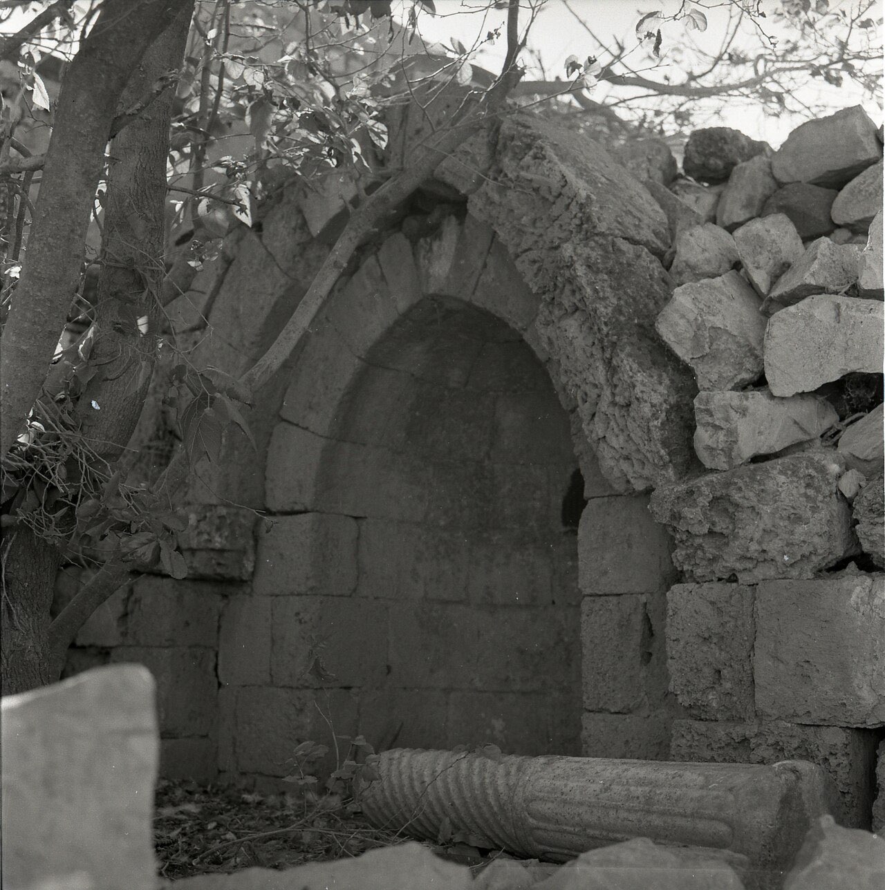 A stone archway in the village of Nein, photographed in black and white, with village buildings and bare hillside behind it.
