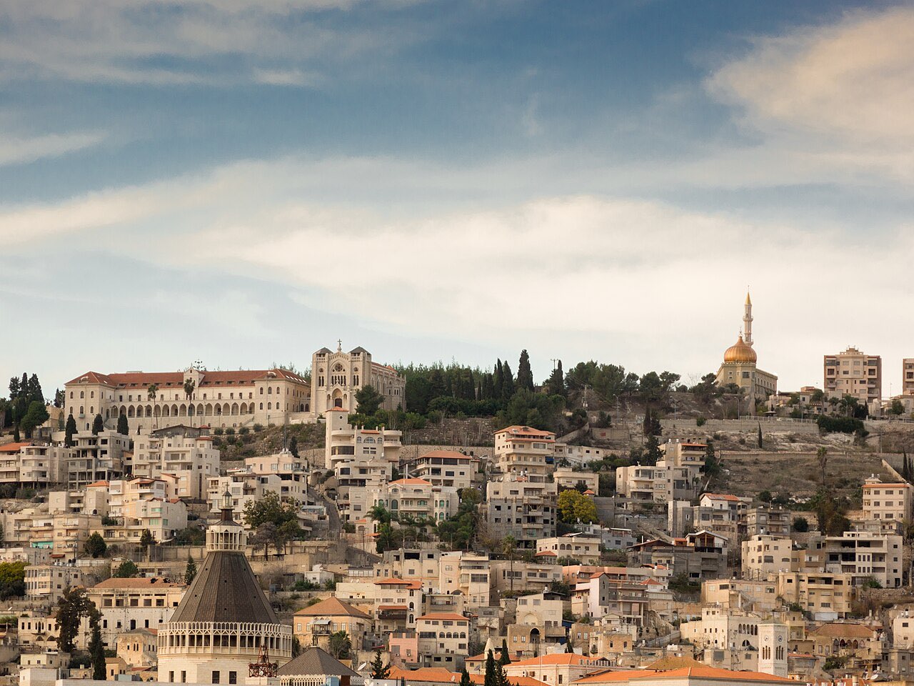 Modern Nazareth climbing its hillside, dense with stone houses and church towers; the dark cone-shaped dome of the Basilica of the Annunciation rises at the lower left.