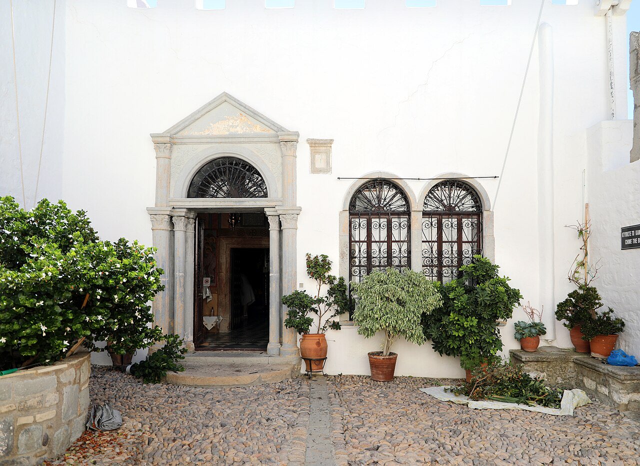 A whitewashed monastery courtyard in Chora on Patmos: a carved stone doorway, arched windows with iron grilles, and potted plants on cobblestones.