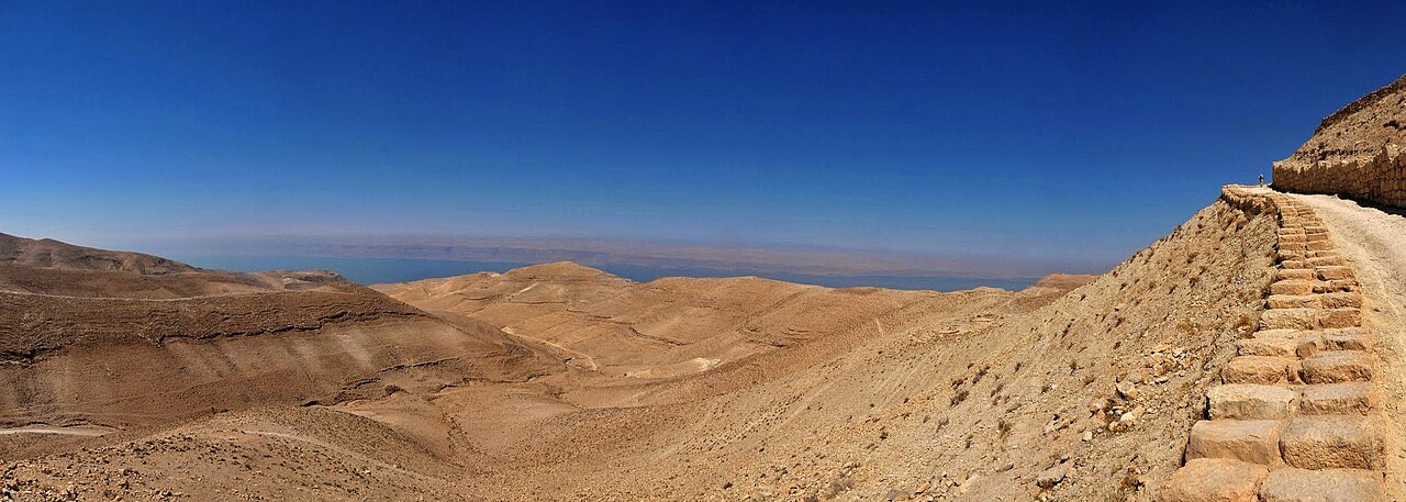 A stone path winding along the barren Perean highlands, with the Dead Sea pale in the haze far below.