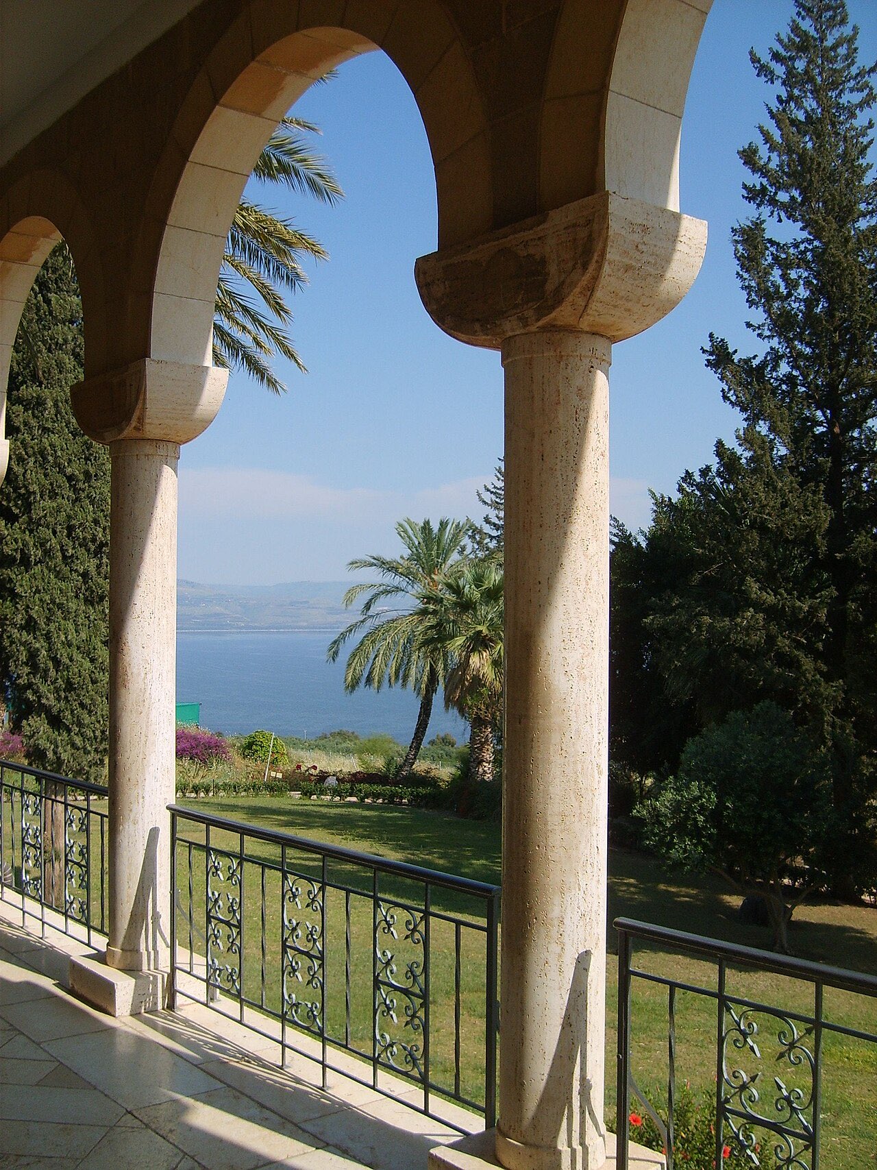The Sea of Galilee seen through the arched stone colonnade of the Church of the Beatitudes, palm trees and gardens below and the hills of the far shore across the blue water.