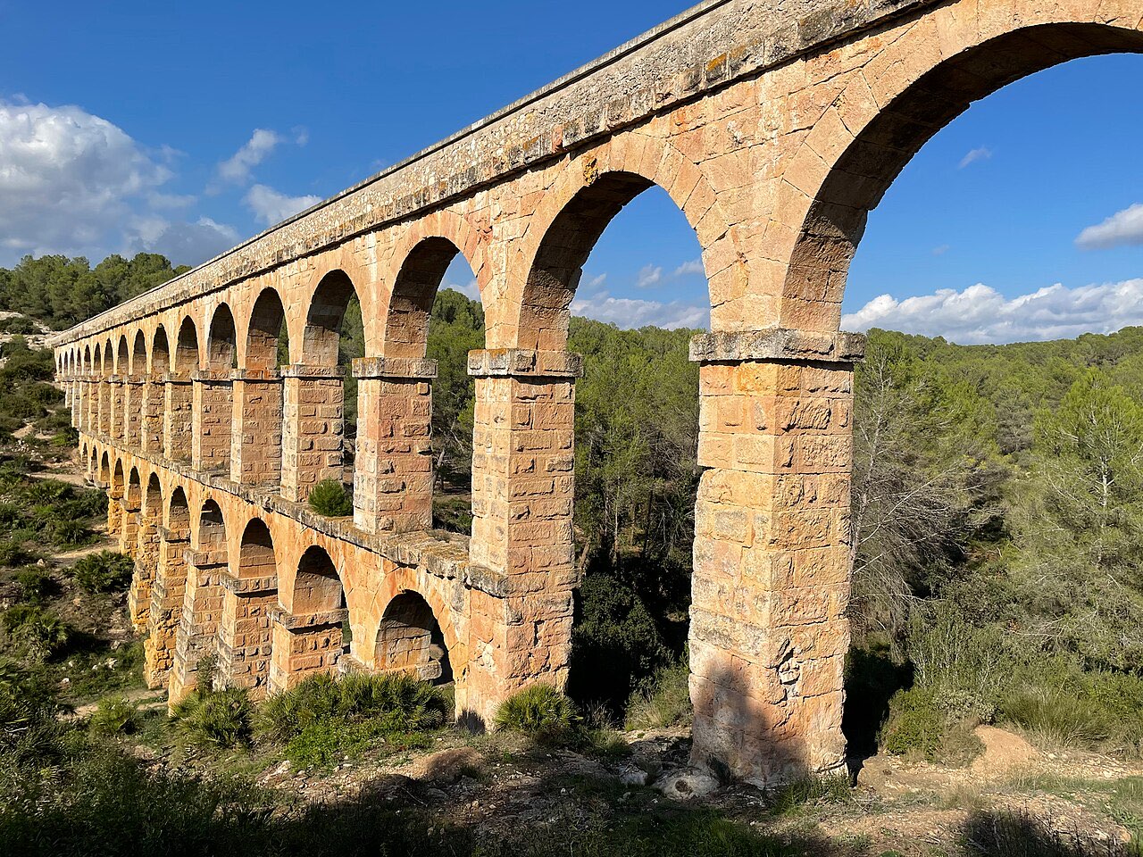 The two tiers of golden stone arches of the Les Ferreres Roman aqueduct near Tarragona, spanning a wooded valley under a blue sky.