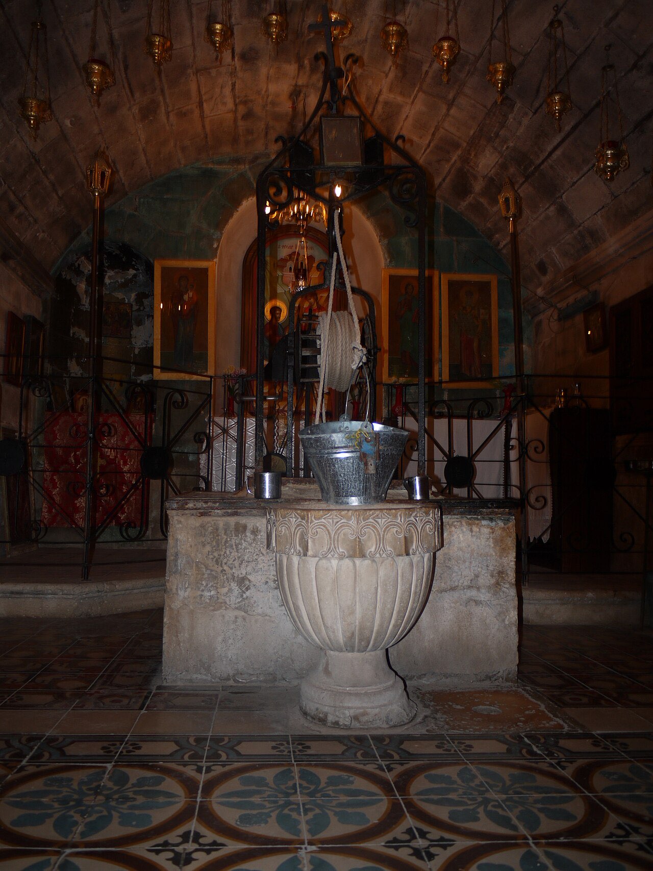 The crypt beneath the Church of St. Photina in Nablus: Jacob's Well, a carved stone wellhead fitted with a winch and metal bucket, surrounded by hanging oil lamps and icons.