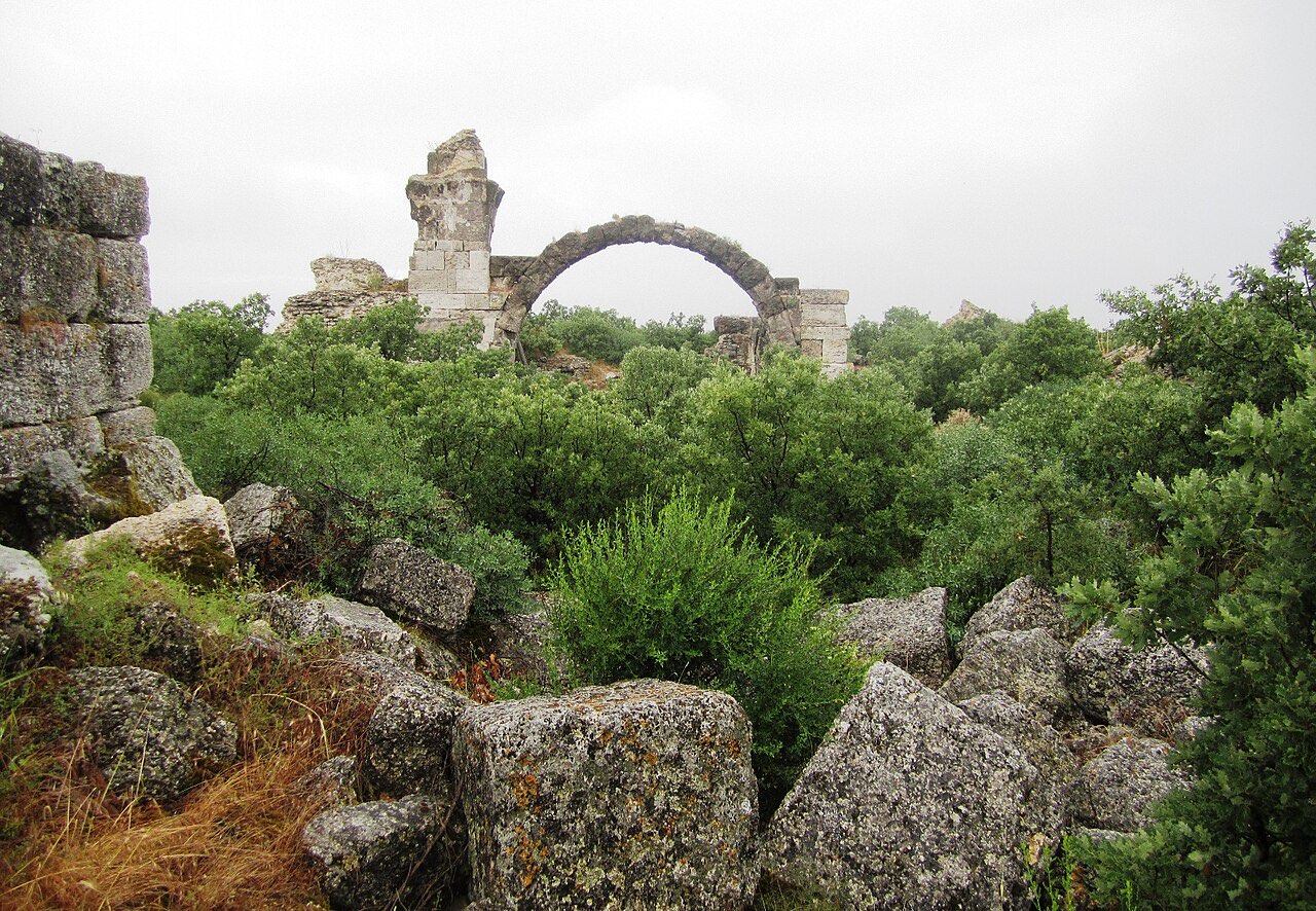 A ruined stone arch of the Roman baths at Alexandria Troas rising above dense green scrub and tumbled blocks.